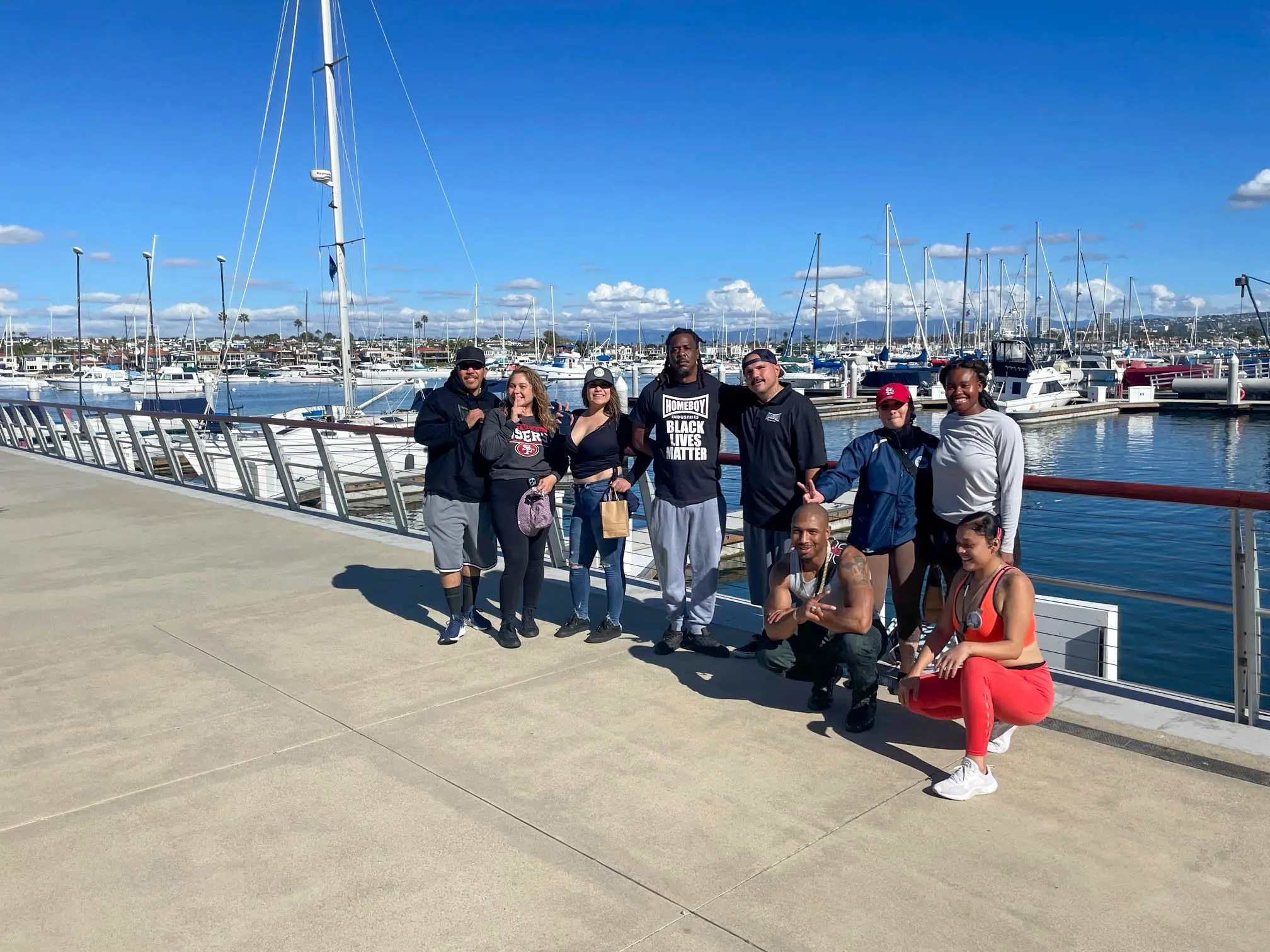A group of participants pose together on a harbor walkway on a sunny day, with sailboats in the marina and blue sky behind them