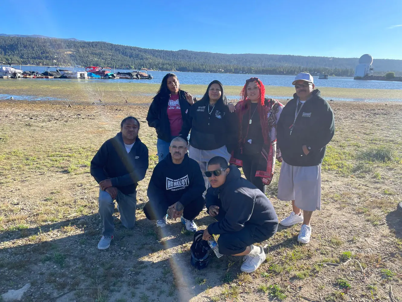 Seven participants and staff pose together on the shore of Big Bear Lake, with boats docked on the water and pine-covered hills behind them