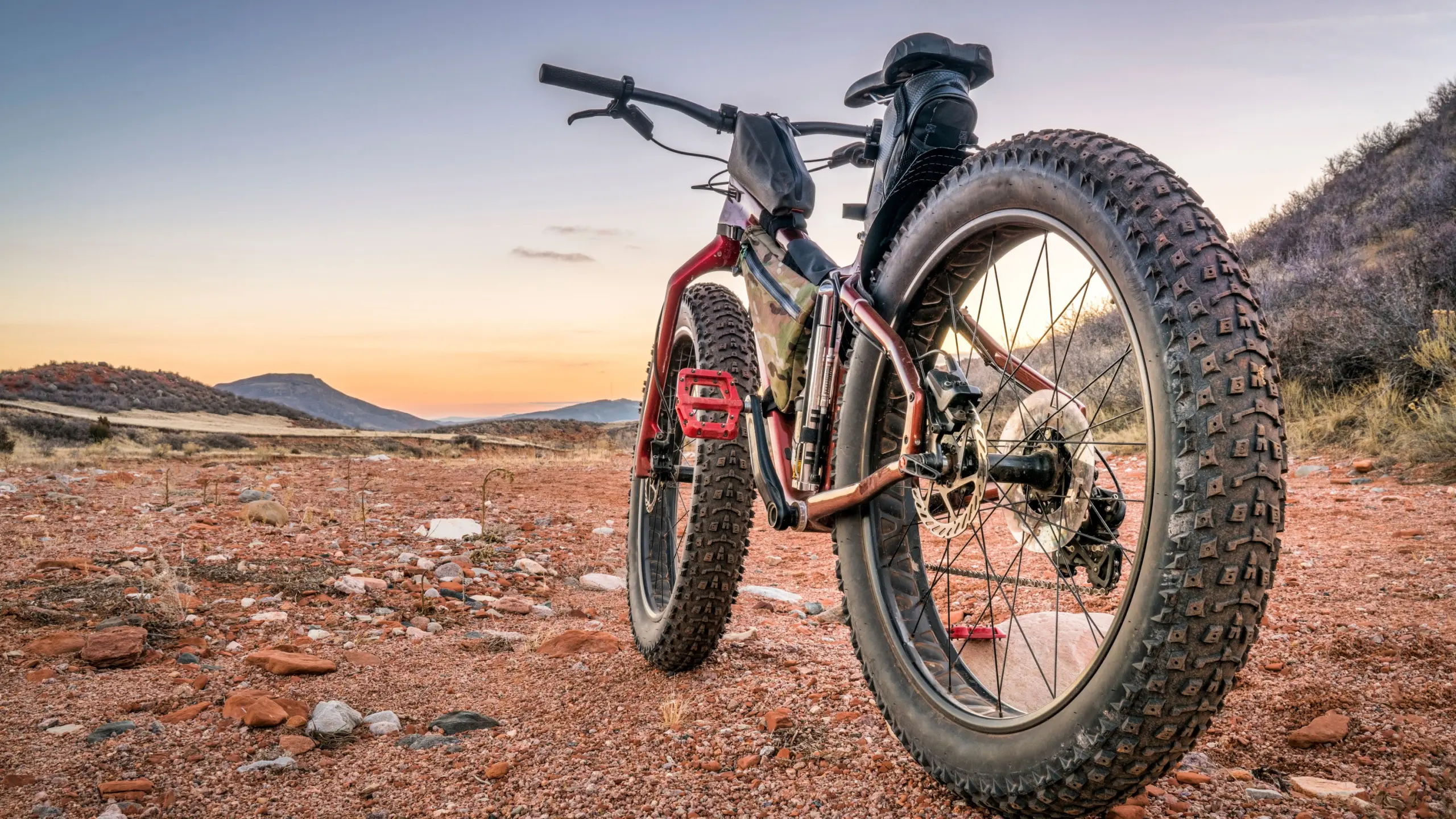 A fat-tire mountain bike with red pedals parked on red rocky desert ground at sunset, with hills on the horizon