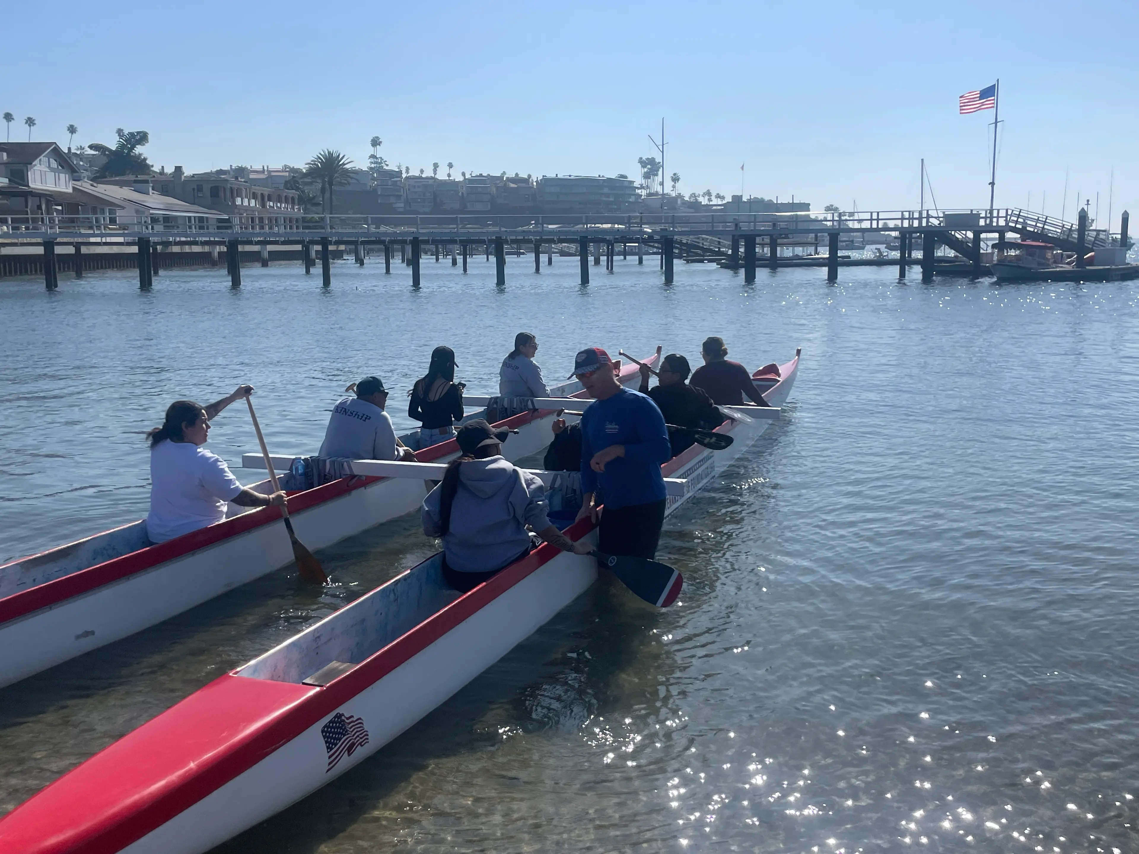 Participants in two red-and-white outrigger canoes get ready to paddle out from a sunny harbor beach, with docks and waterfront homes behind them