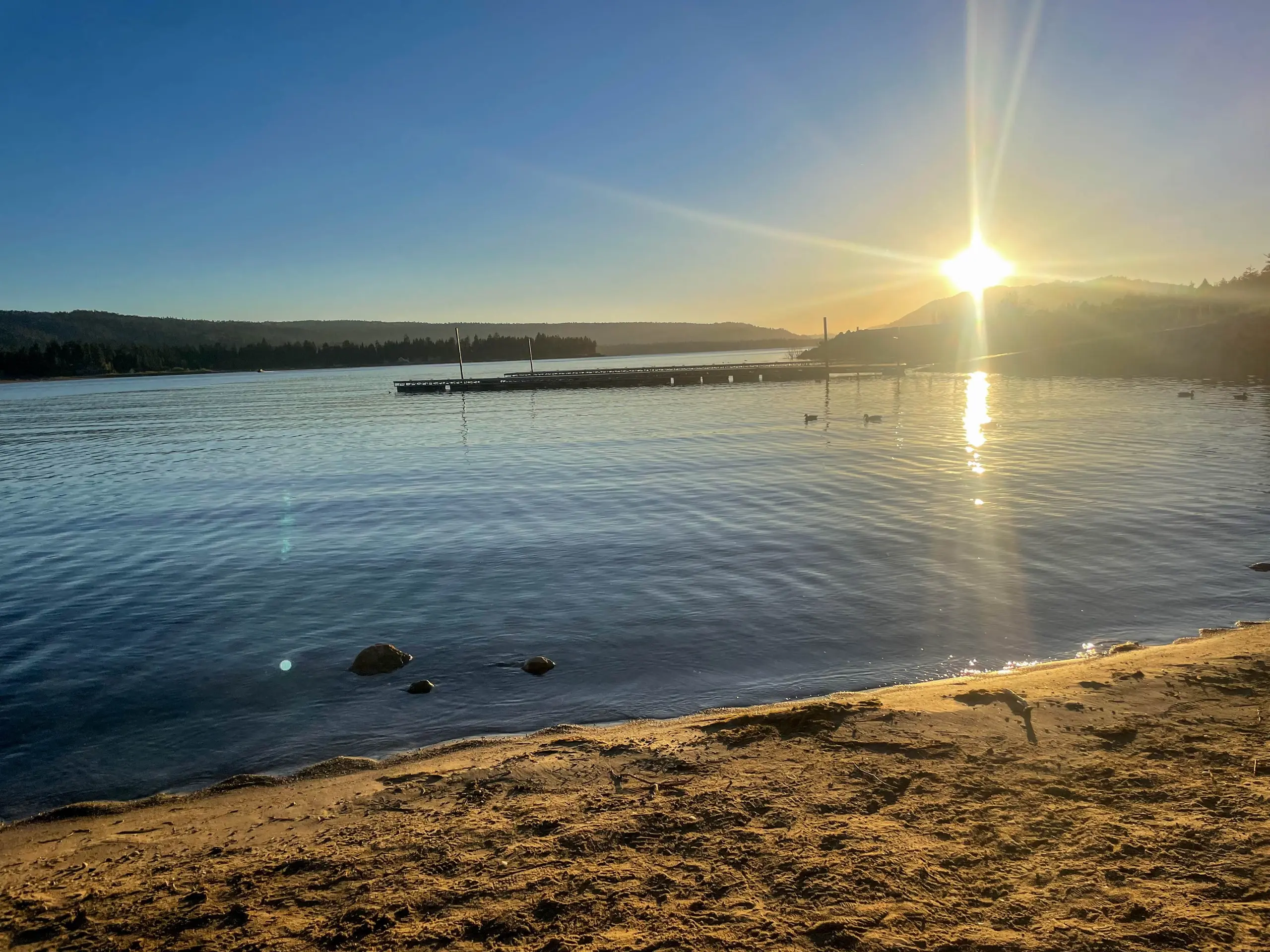 The sun sets over Big Bear Lake, with a boat dock and ducks on the calm water and a sandy shoreline in the foreground