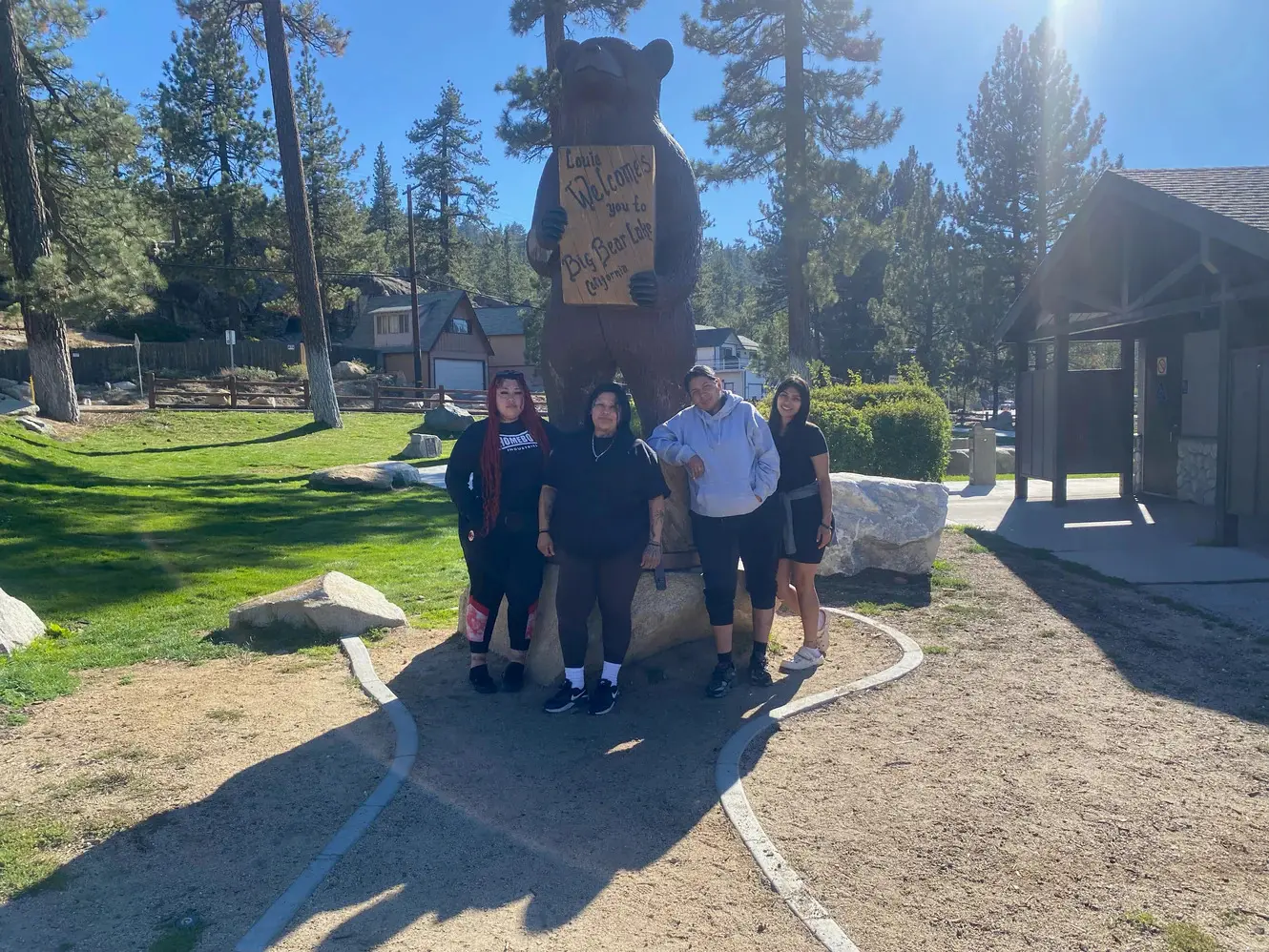 Four participants pose in front of the carved wooden bear holding a Welcome to Big Bear Lake sign, on a green lawn in the pines