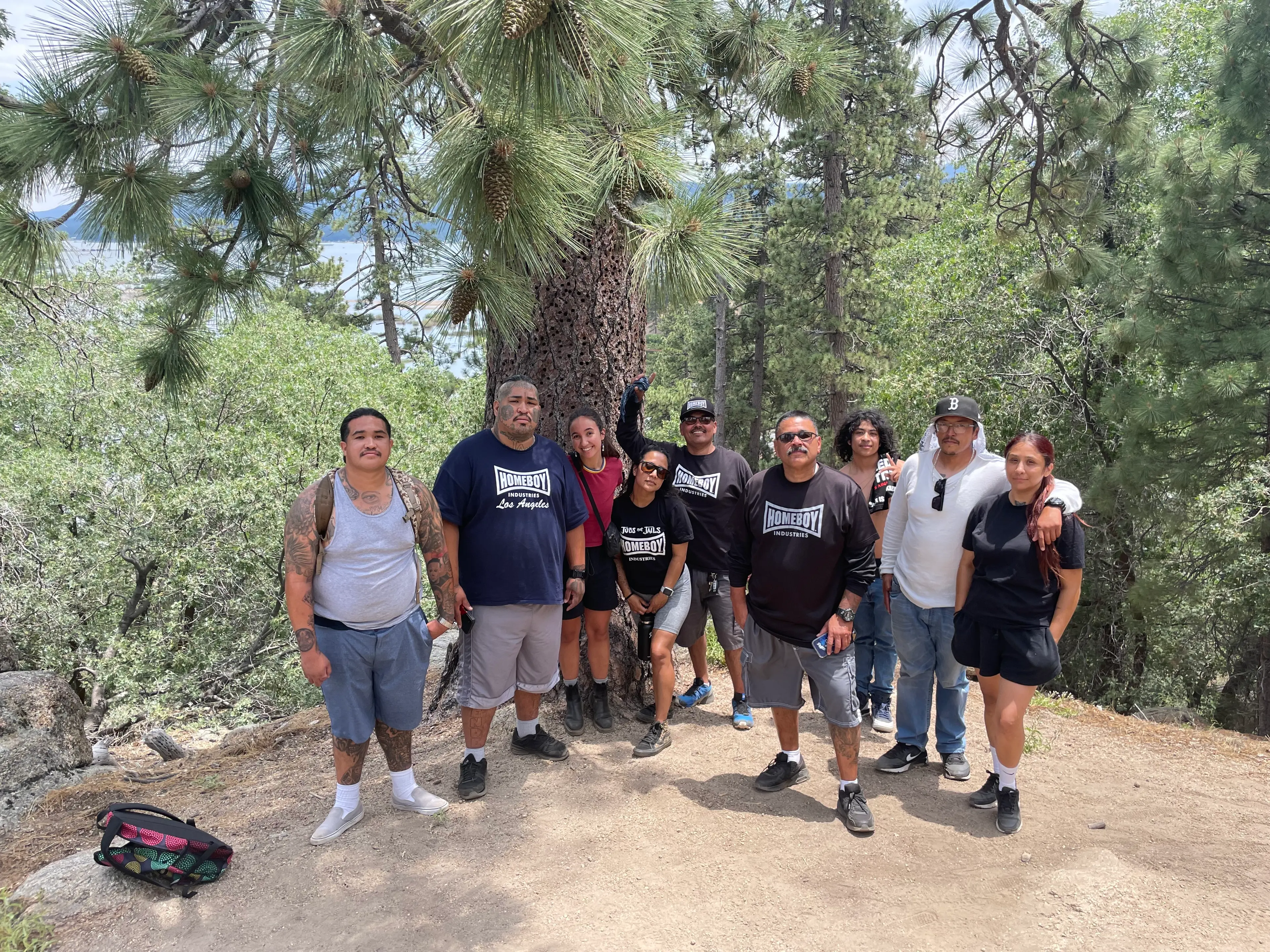 A group of Outside the City participants standing together on a forest trail under tall pines, with Big Bear Lake behind them