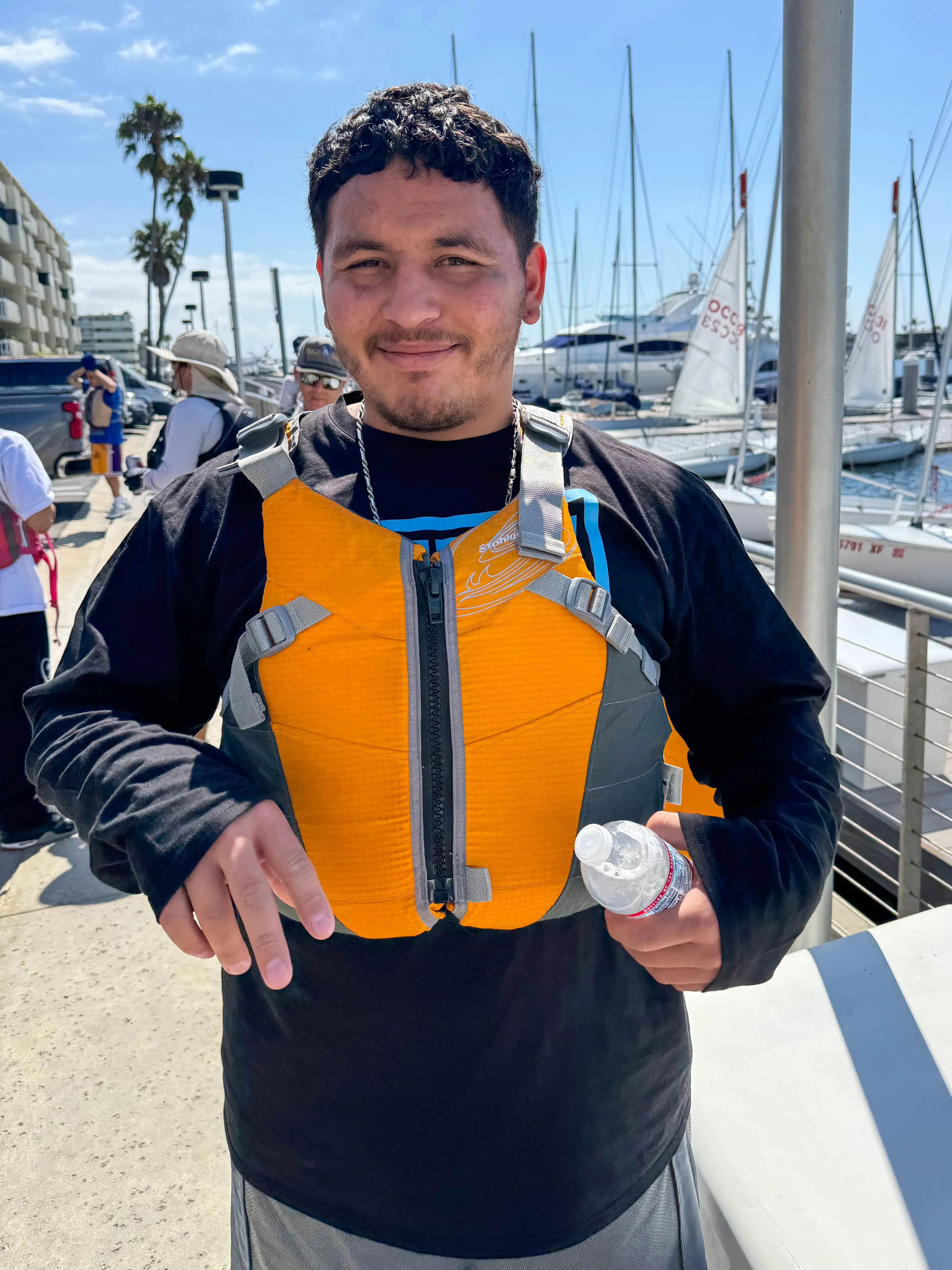 A smiling participant in an orange life vest holds a water bottle on the Marina Park dock in Newport Beach, with sailboats moored behind him