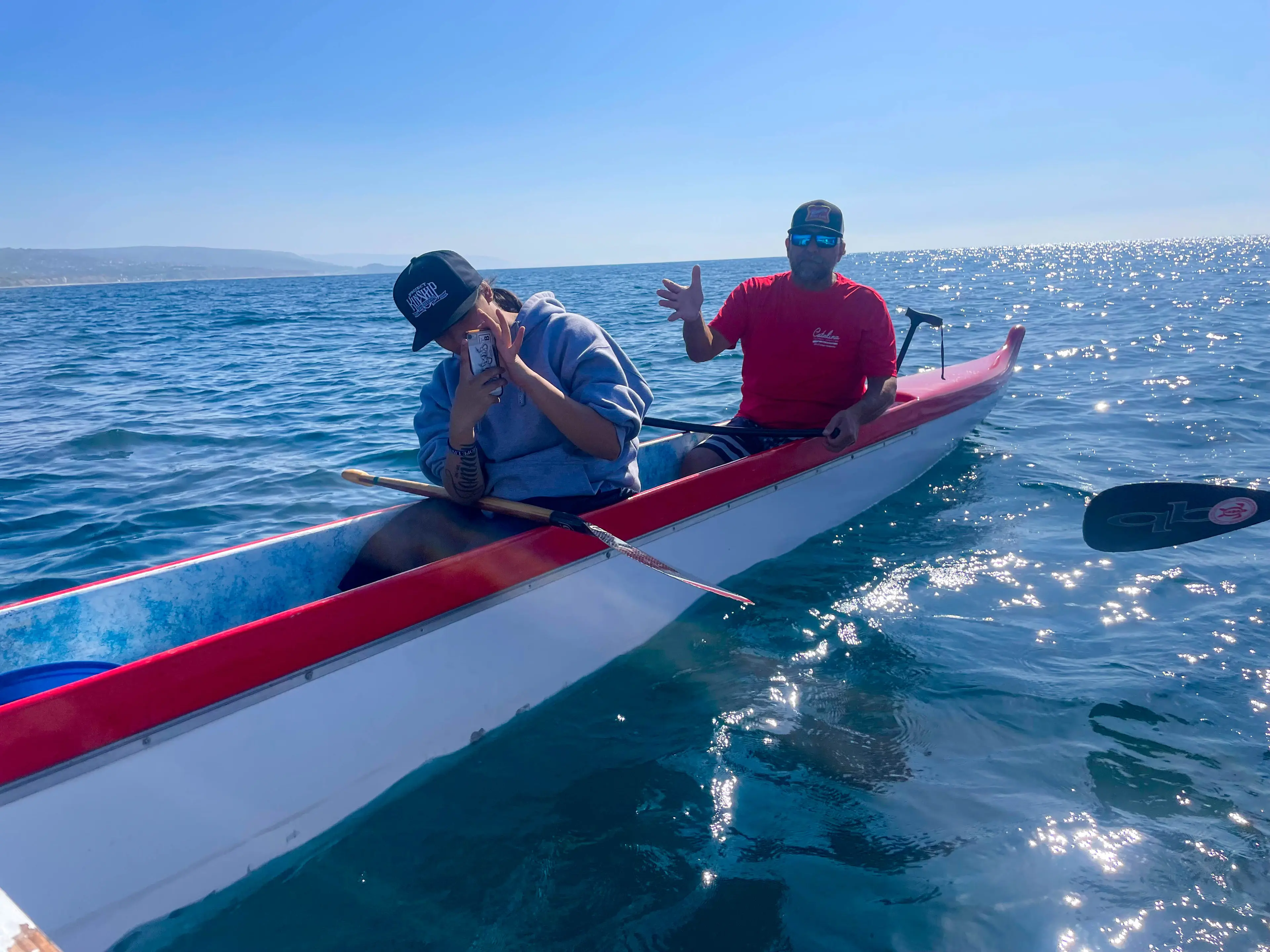 Two people in a red-and-white outrigger canoe on the open ocean off Newport Beach, with the coastline behind them