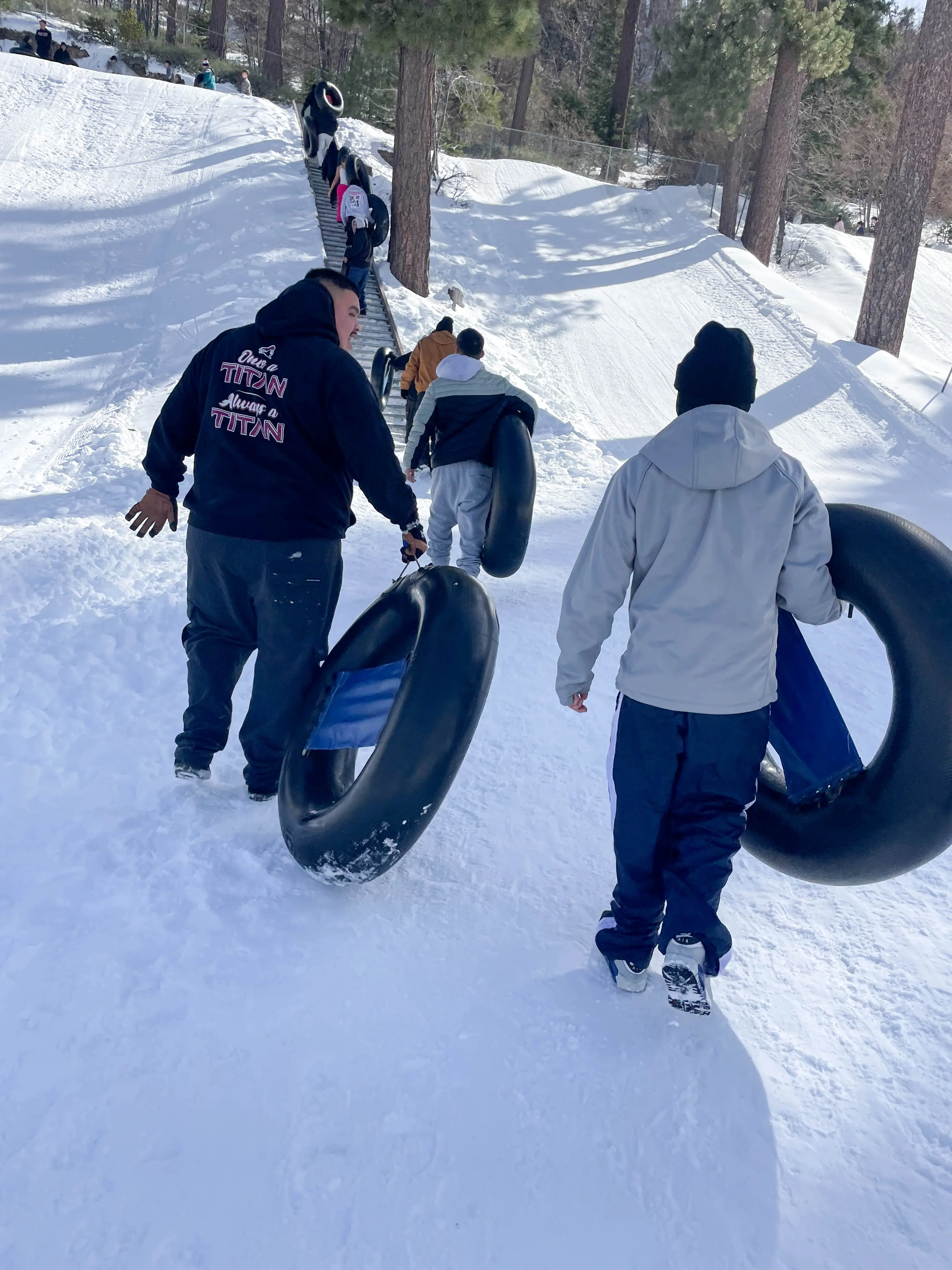 Participants in winter jackets haul black snow tubes up a packed-snow slope at Running Springs, with a line of people climbing the stairs behind them