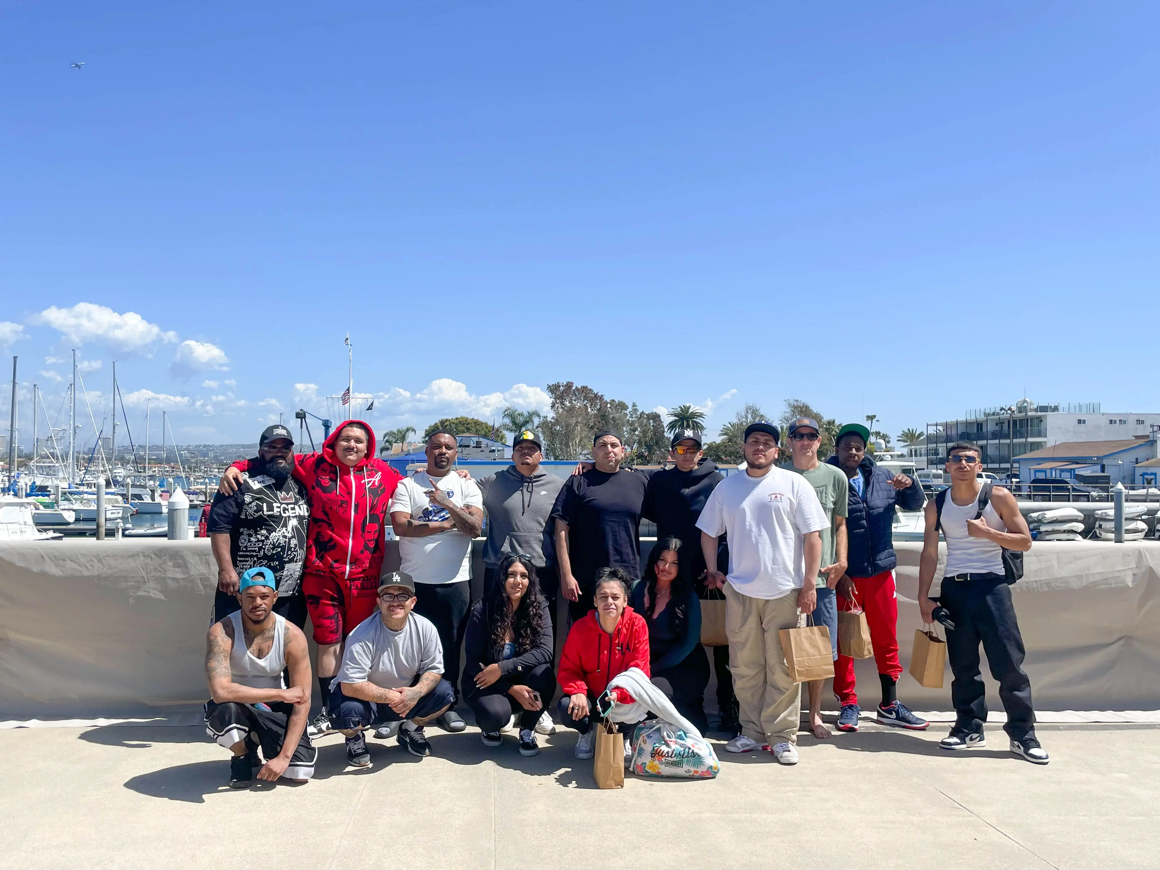 A large group of Outside the City participants gathered on the boardwalk at Marina Park in Newport Beach, with the harbor behind them