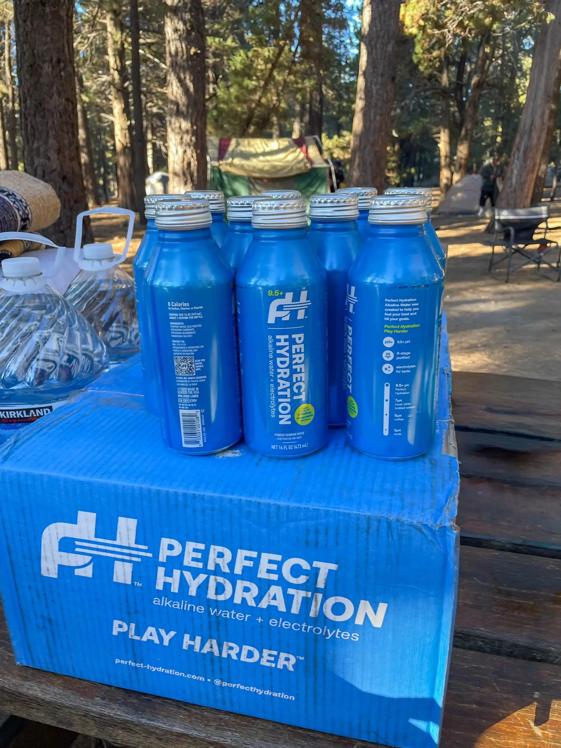 Cases of Perfect Hydration alkaline water and gallon jugs stacked on a picnic table at a Big Bear campsite, with tents among the pine trees behind