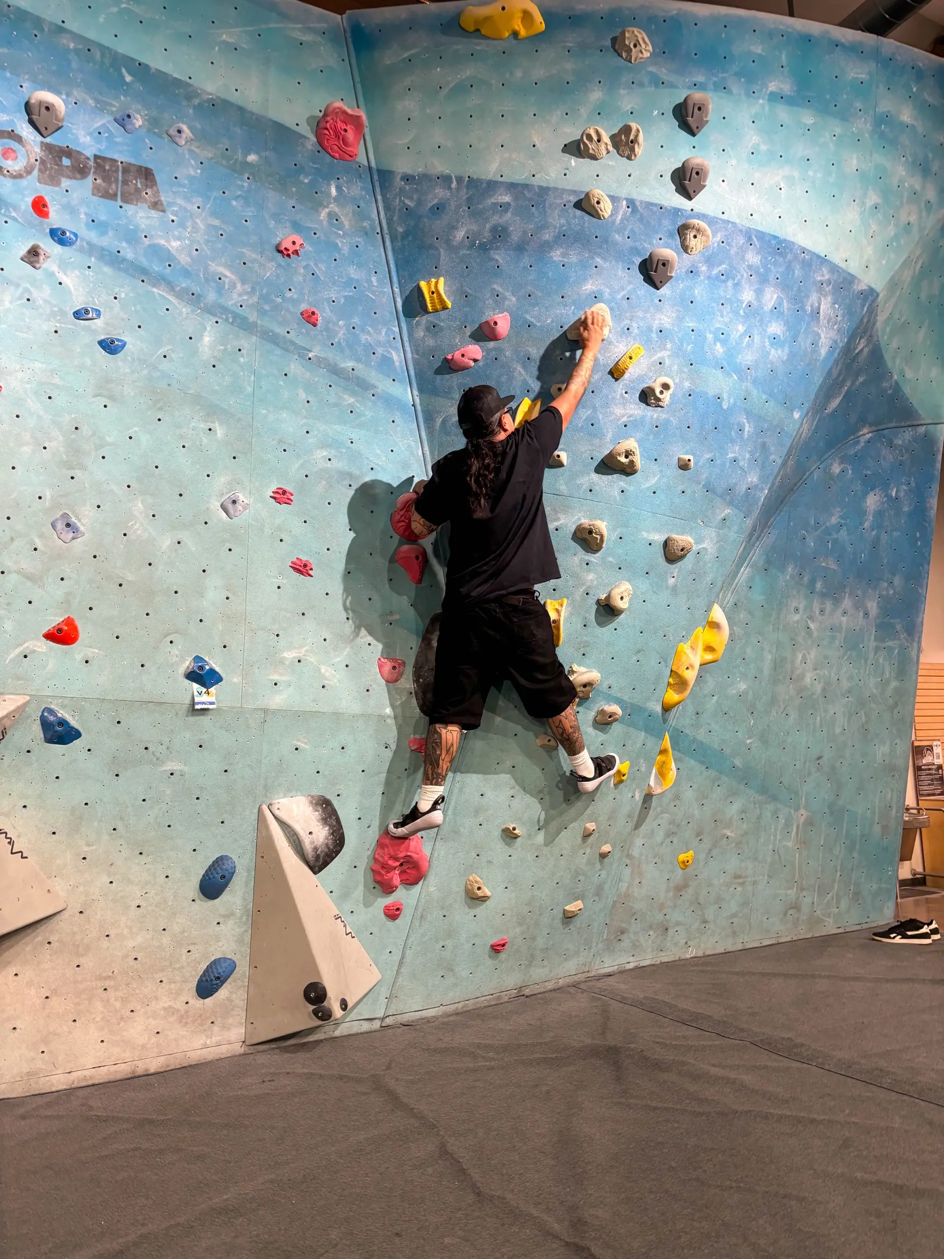A climber in a black cap and black shirt, seen from behind, reaches for a hold on a teal indoor bouldering wall at Sender One
