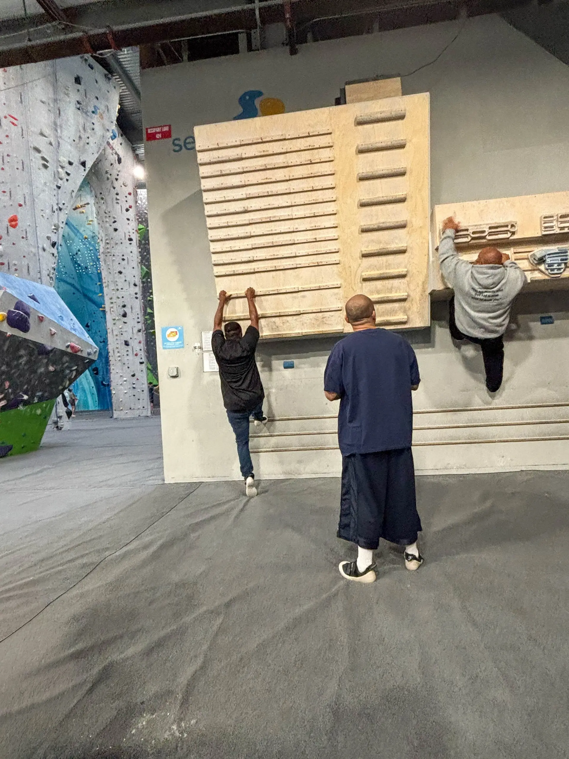 Participants hang from rungs on a wooden training wall at an indoor climbing gym while another participant watches
