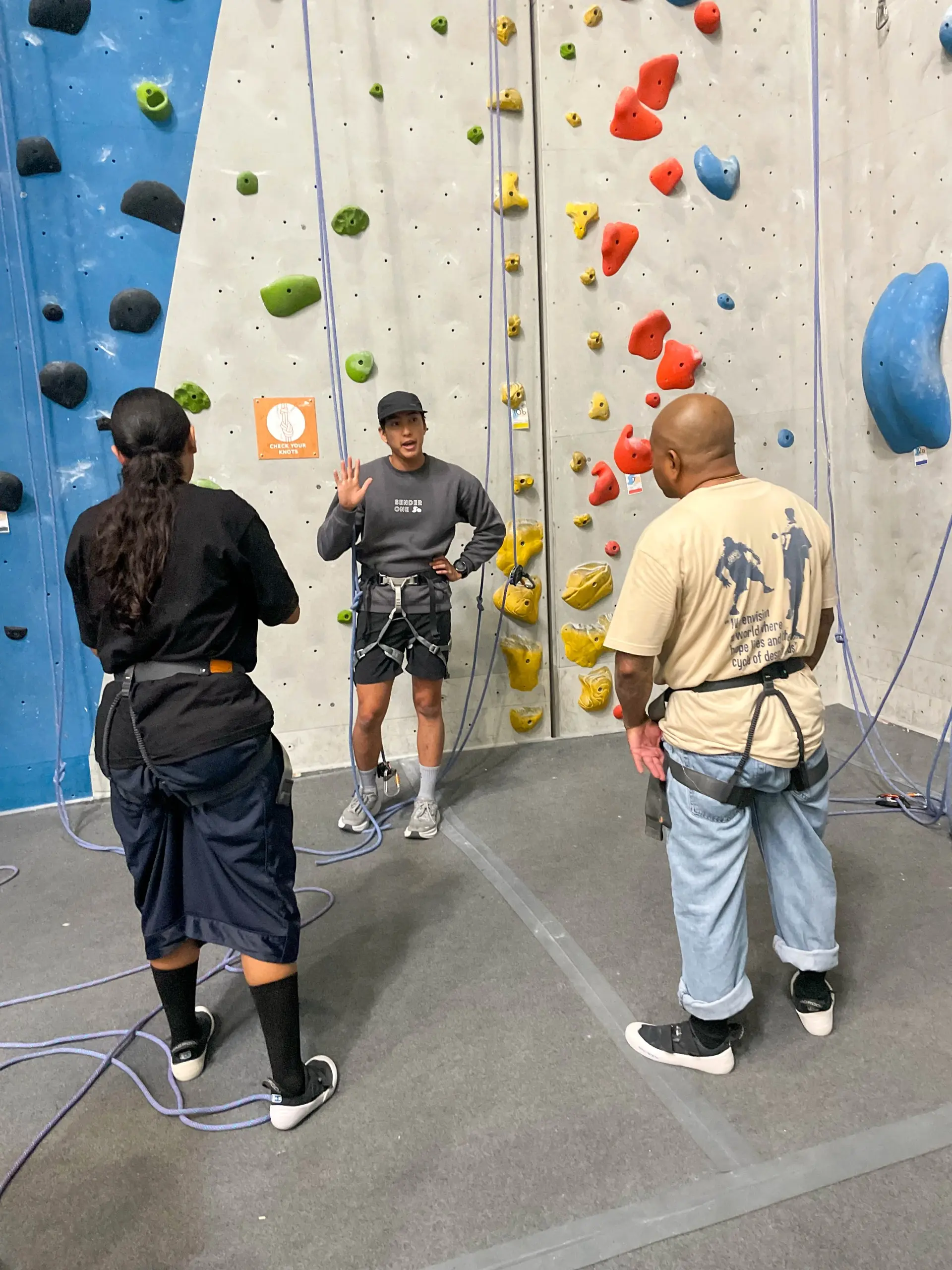 A climbing instructor in a harness explains safety steps to two participants in harnesses, in front of an indoor climbing wall with ropes and colorful holds