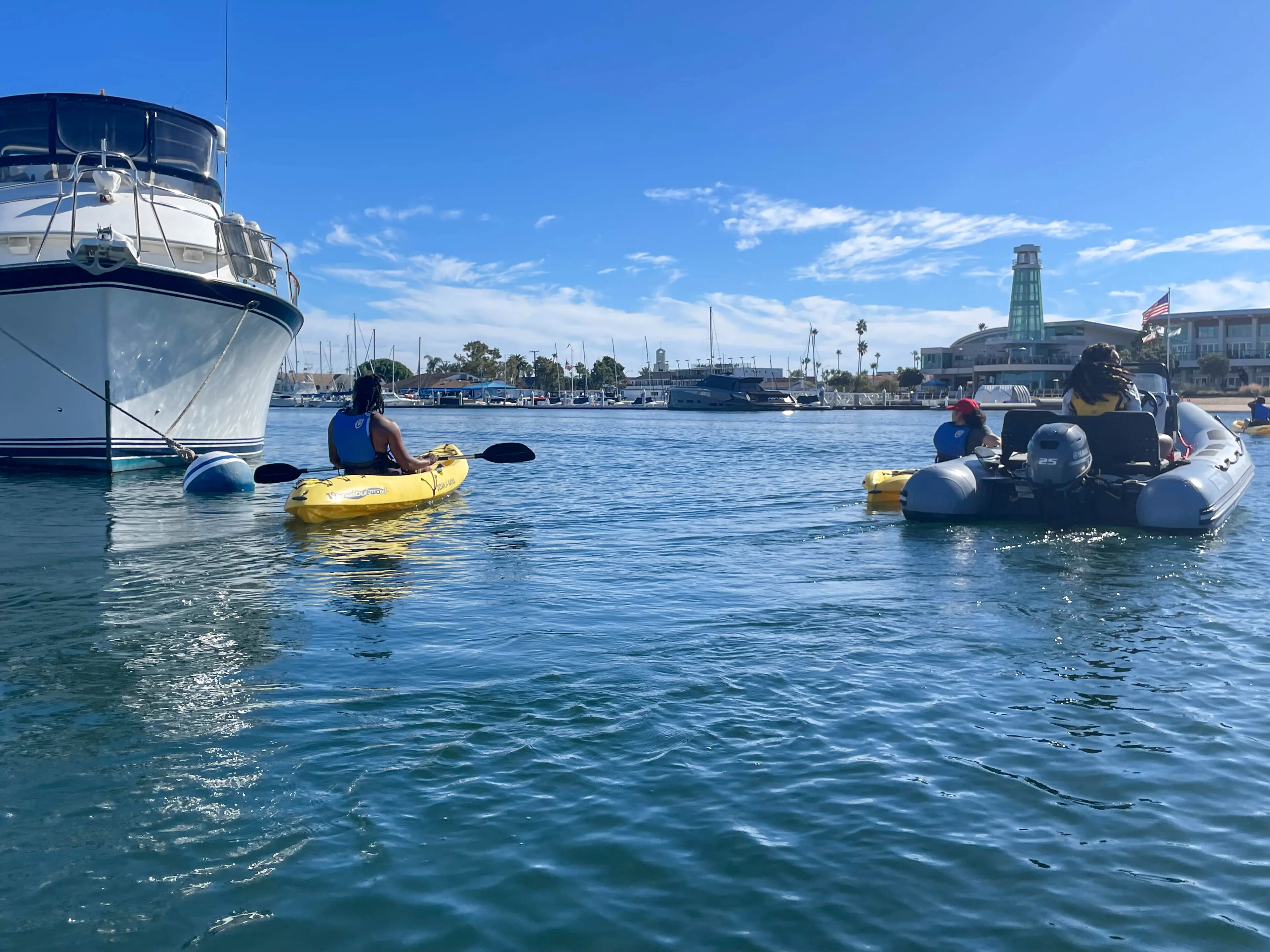 A participant paddling a yellow kayak past a moored yacht on Newport Harbor, with others in an inflatable boat nearby and the Marina Park lighthouse across the water