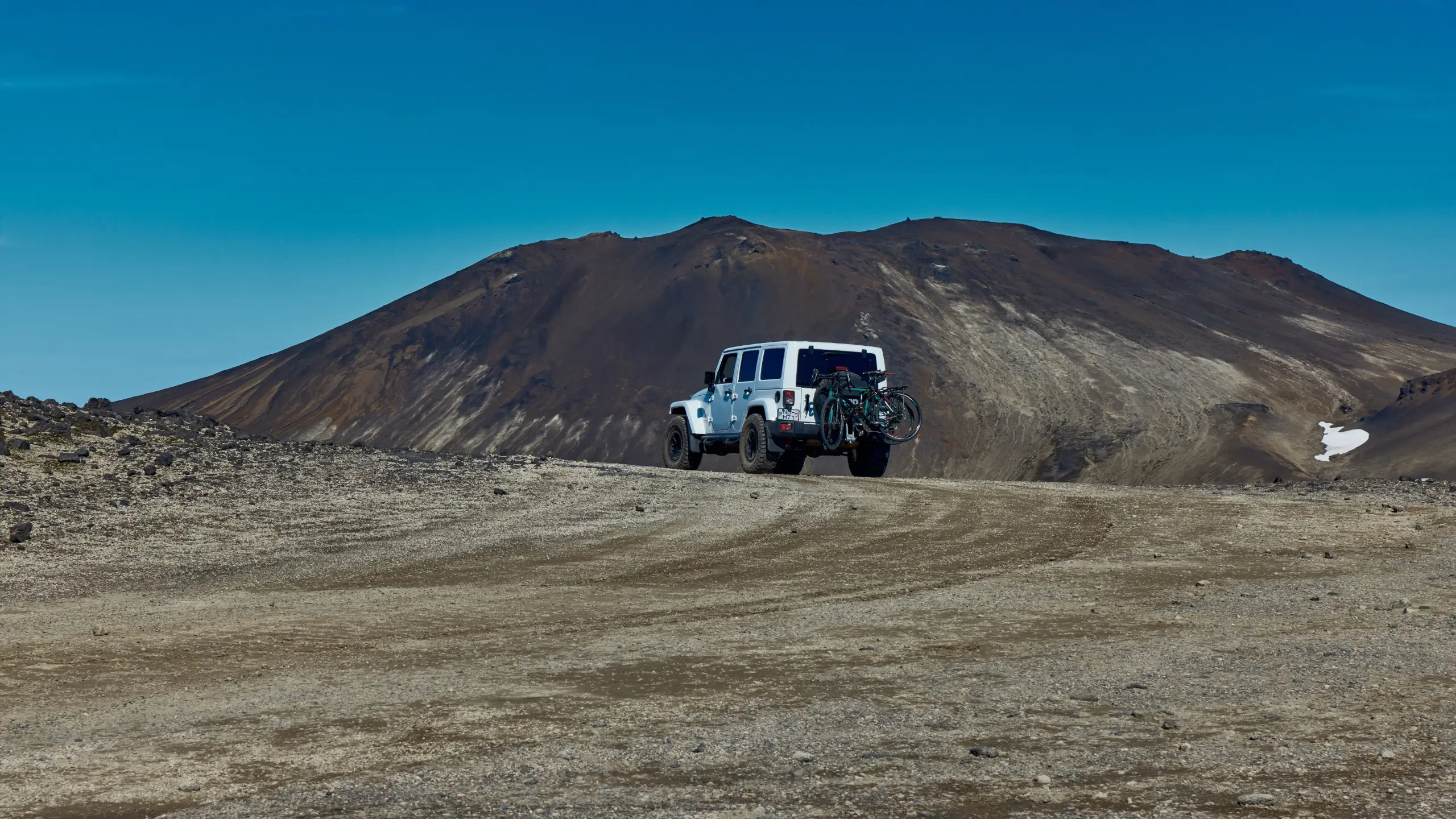 A white Jeep Wrangler with two bikes on a rear rack drives a gravel track toward a dark volcanic mountain in the Icelandic Highlands, under a clear blue sky