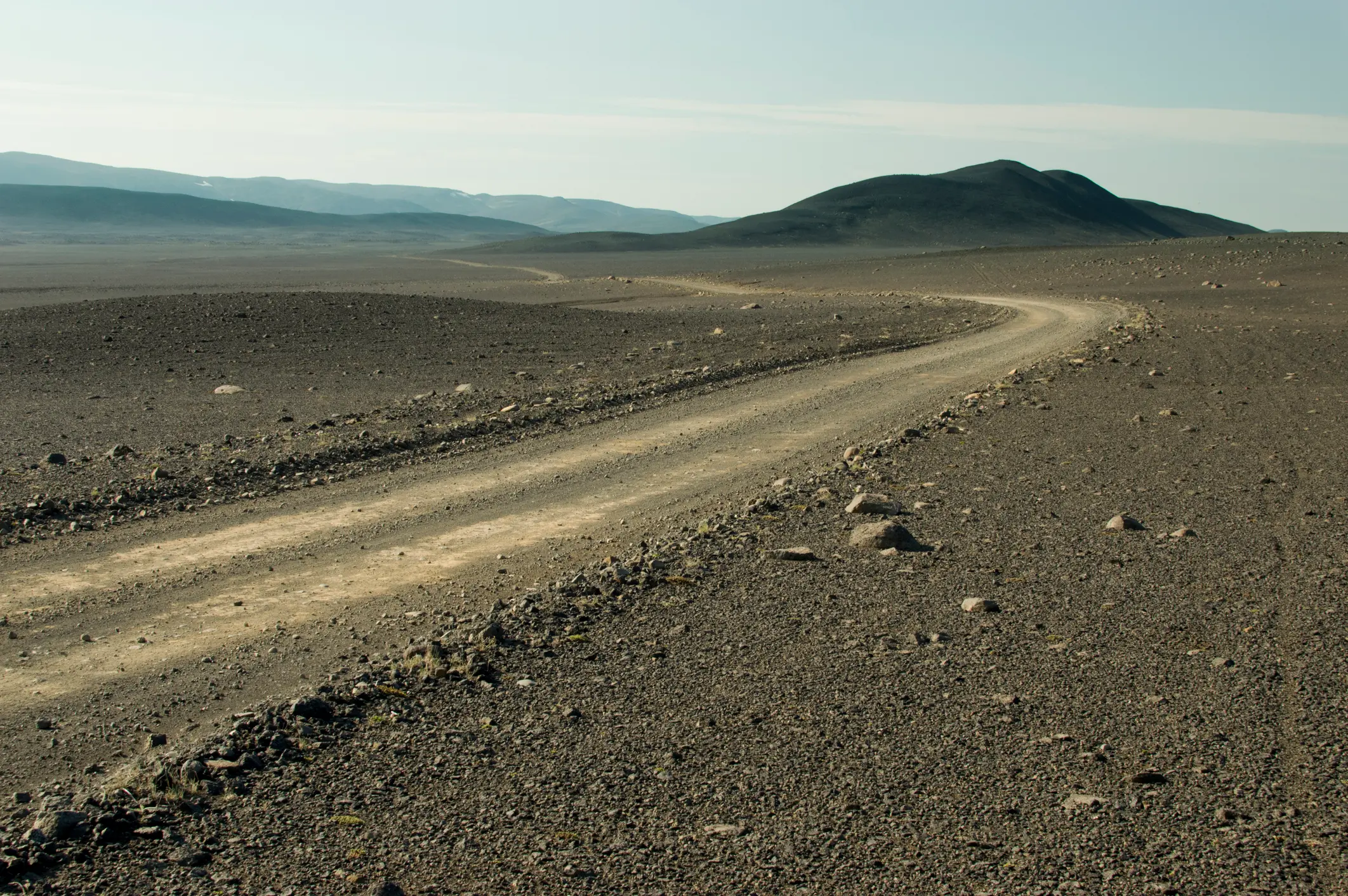 A gravel track winds across a barren stony plain toward dark volcanic hills in the Icelandic Highlands