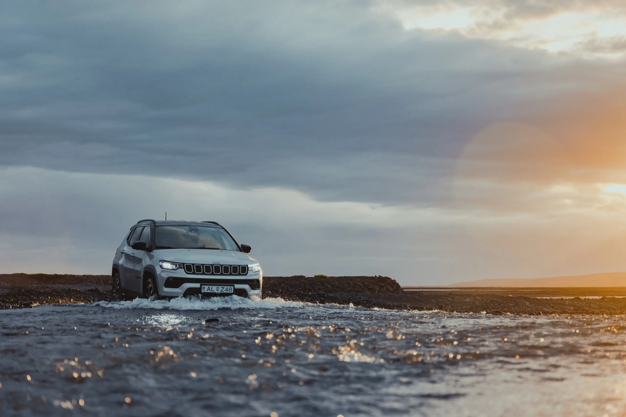 A white Jeep Compass fords a shallow river over black gravel in Iceland, under storm clouds lit by the setting sun