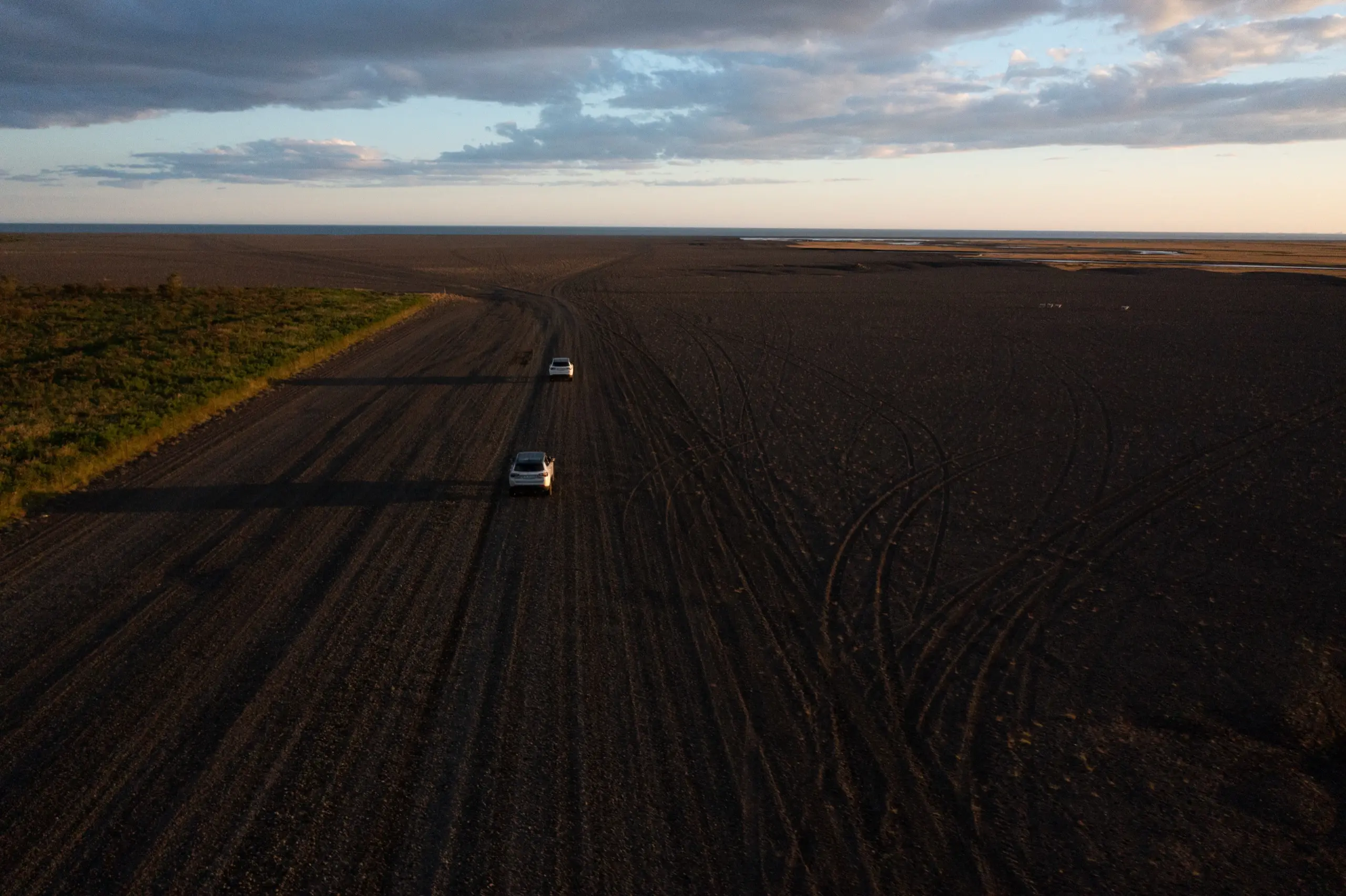 Two white SUVs drive a wide gravel road across a vast dark volcanic plain in Iceland, toward the ocean horizon at sunset