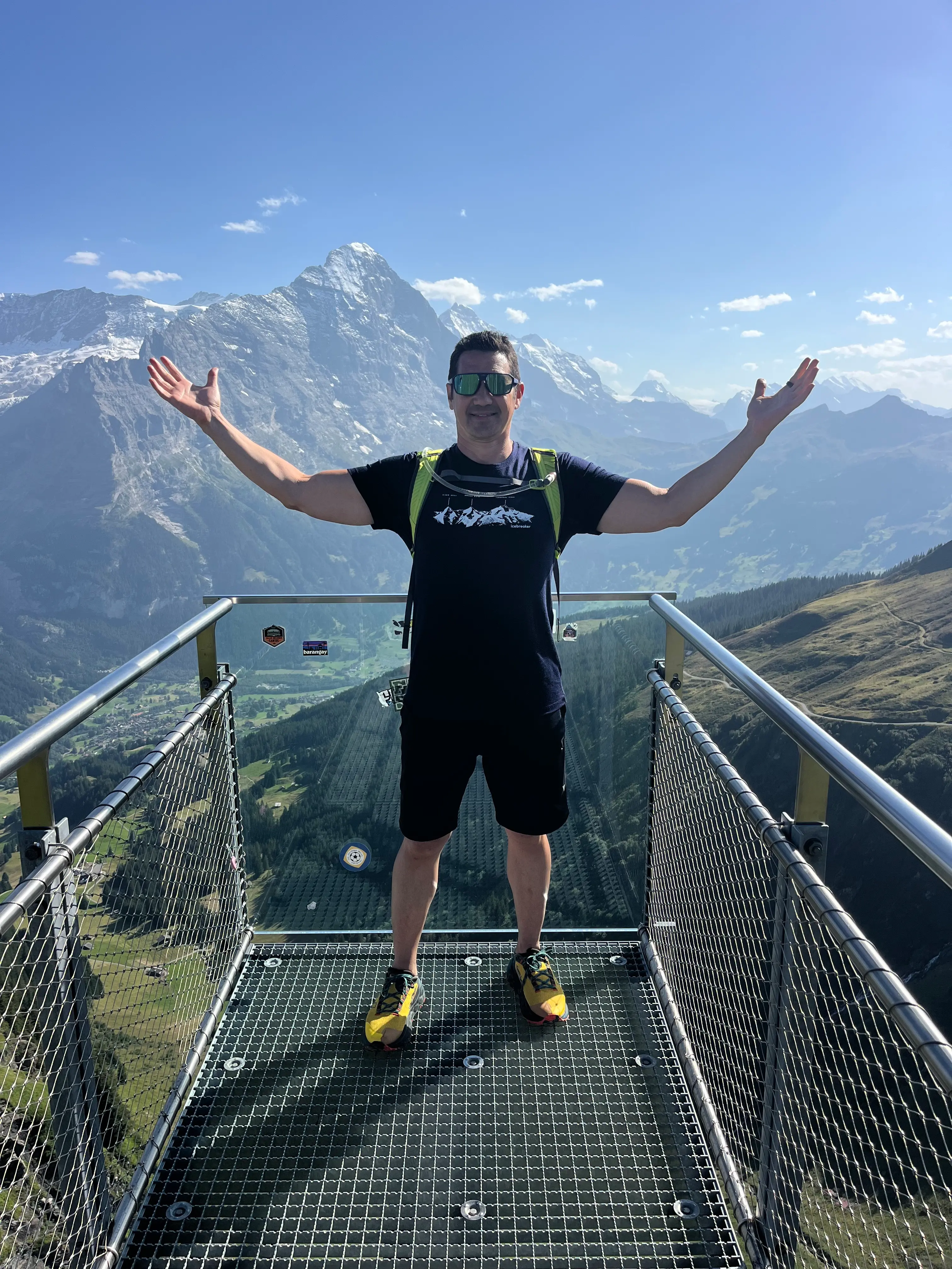 A hiker stands with arms raised on a railed overlook platform, with mountain peaks behind