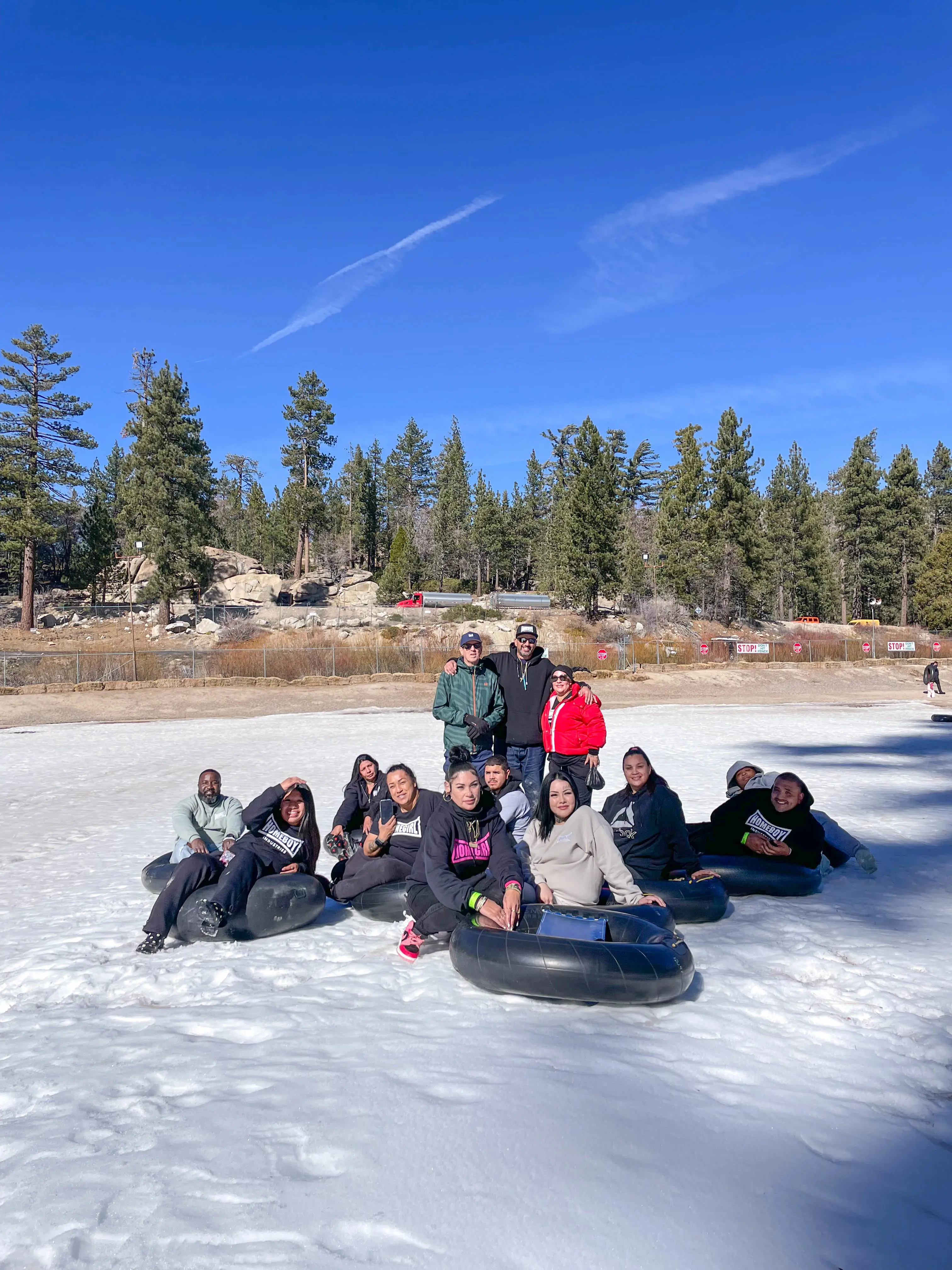 A large group sits together on snow tubes in Running Springs under a clear blue sky, with pine trees behind