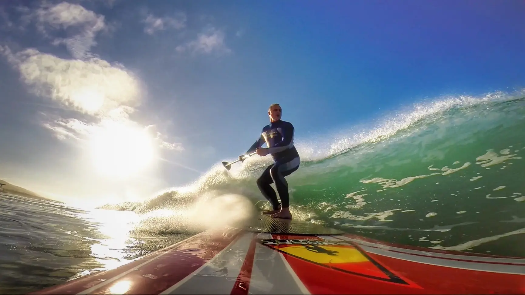 A paddleboarder in a wetsuit rides a glassy green wave toward the camera, paddle in hand, with the sun low in a blue sky behind