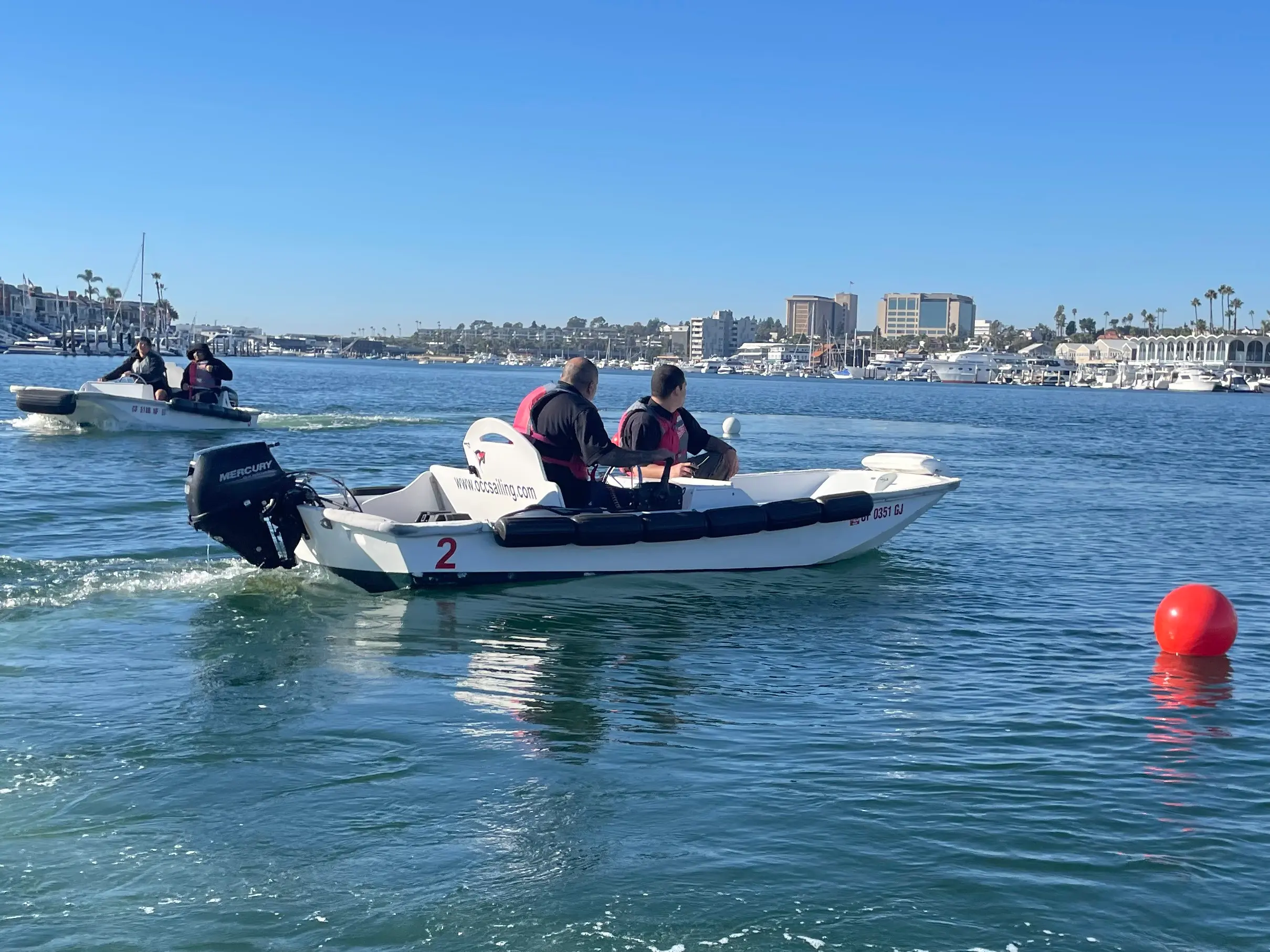 Participants in life vests drive small white training powerboats across Newport Harbor past a red buoy, with the waterfront behind