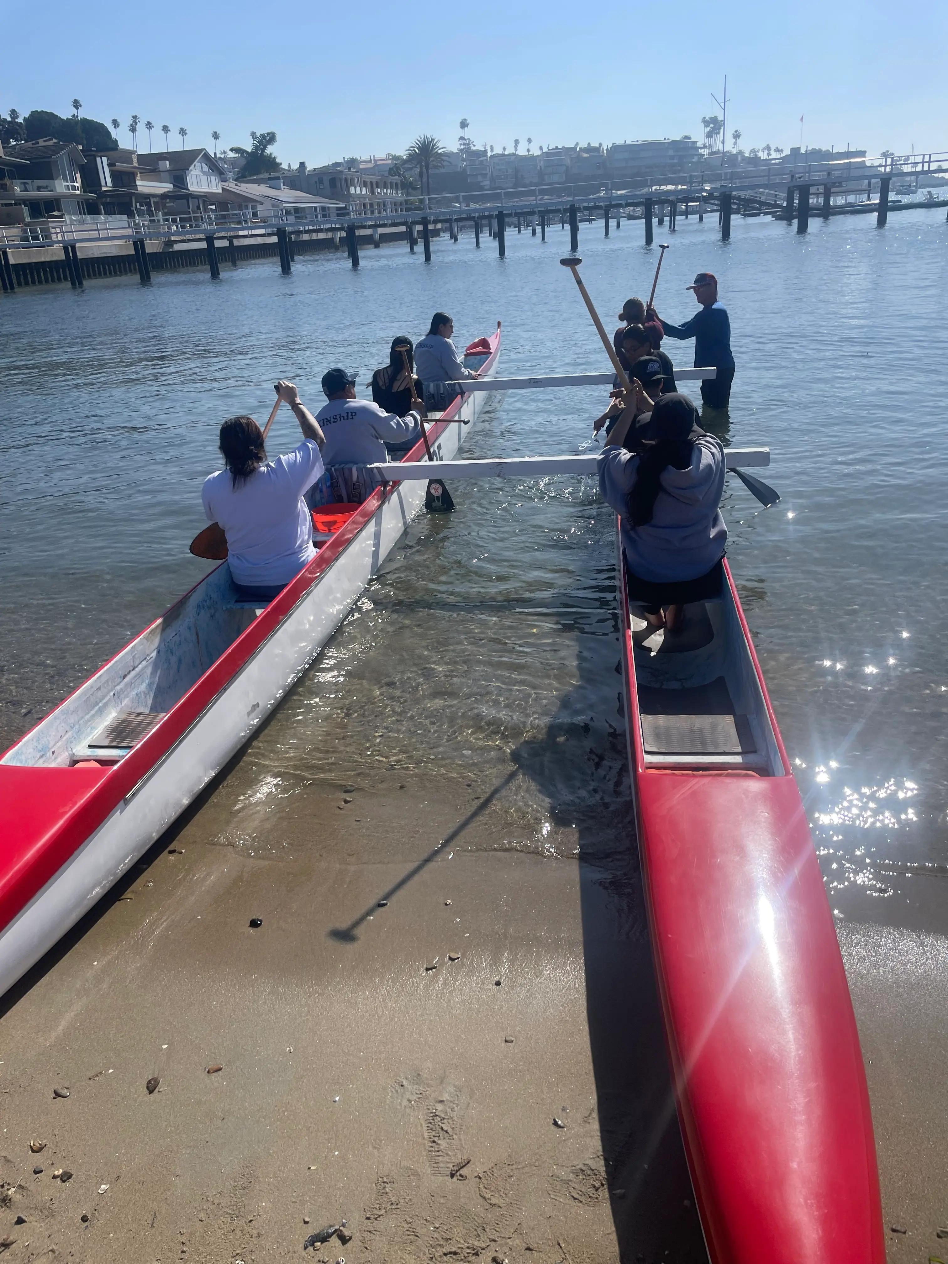 Paddlers climb into two red outrigger canoes at the edge of a calm harbor beach, with docks and waterfront homes behind