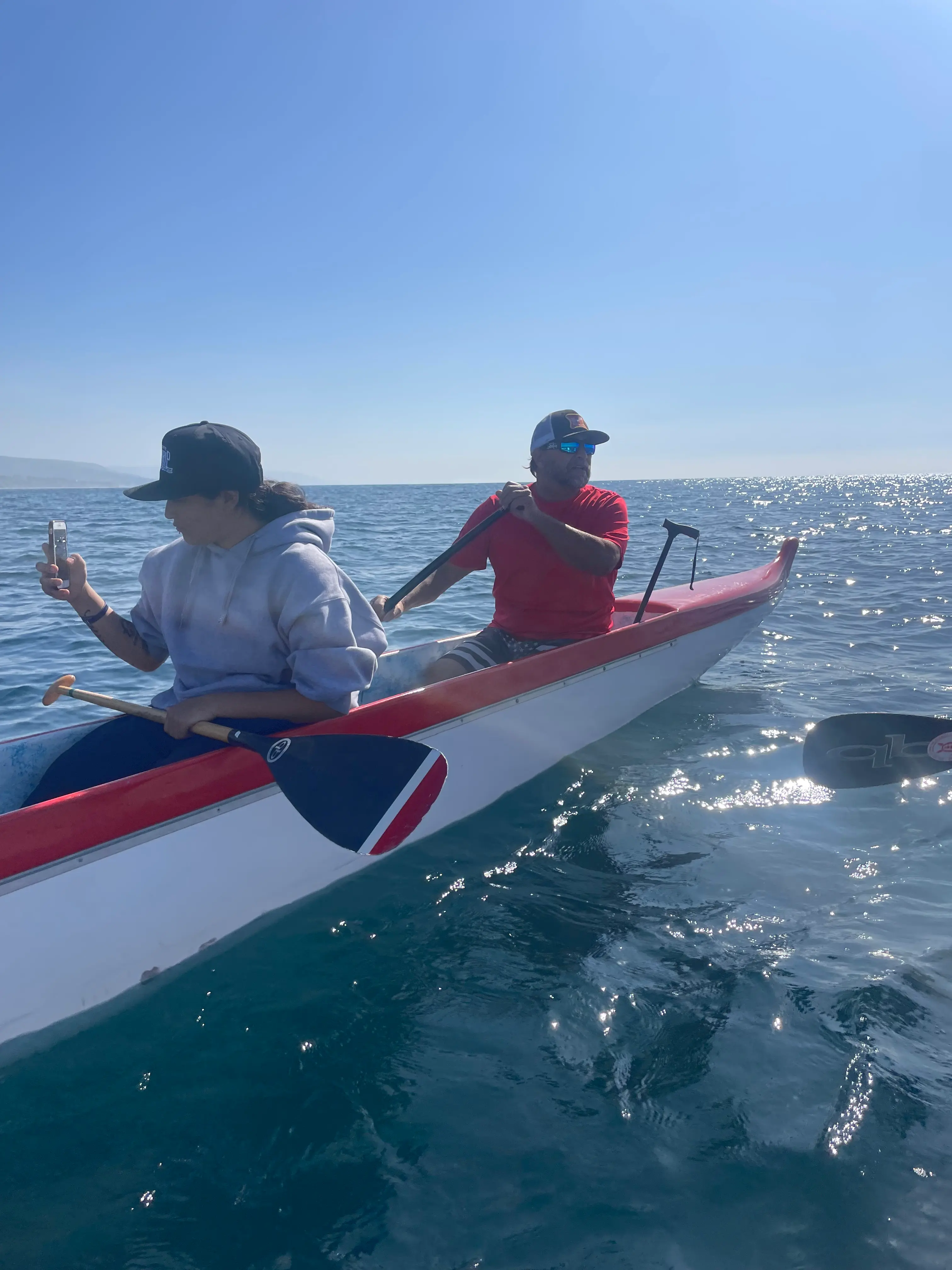 Two paddlers in a red-and-white outrigger canoe on a calm, sunlit ocean